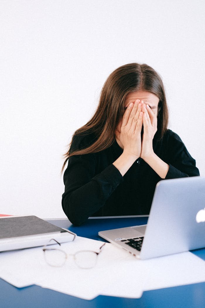 A woman covers her face with hands in stress sitting at an office with a laptop and papers.